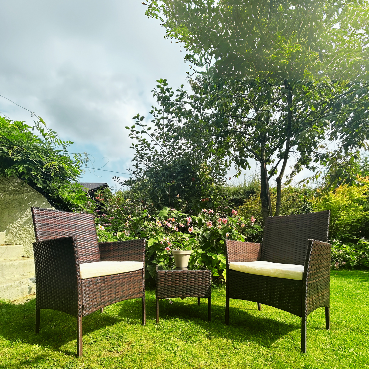 Garden furniture set with chairs and a table on a grassy area with trees and flowers in the background.