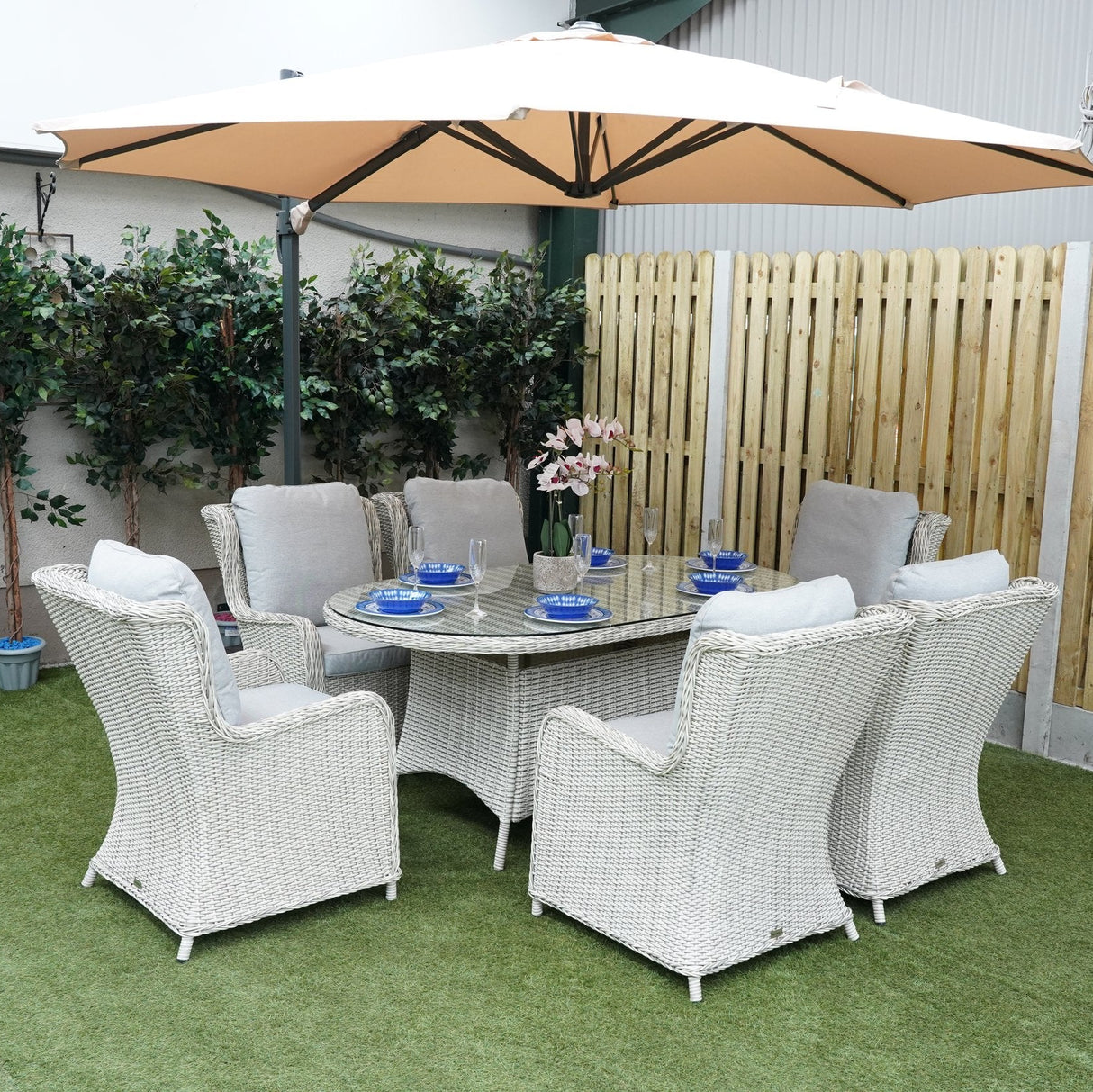 A white rattan dining set with six chairs and an oval table with a glass top, set on a garden patio with a large umbrella overhead.