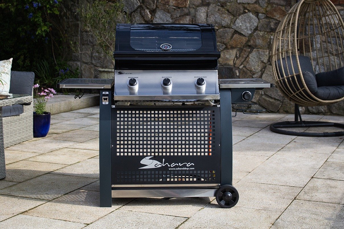 A black Sahara S375 3 Burner Gas BBQ with side burner, placed on a tiled outdoor area with a rock wall and a chair in the background.