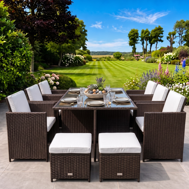 An outdoor 11-piece wicker dining set on a stone patio, featuring a rectangular glass-top table, six armchairs with white cushions, and four matching ottomans, set against a lush green garden background.