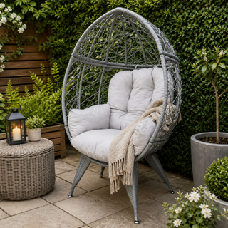 Gray wicker egg-shaped chair with white cushions on a patio, surrounded by plants and a lantern.