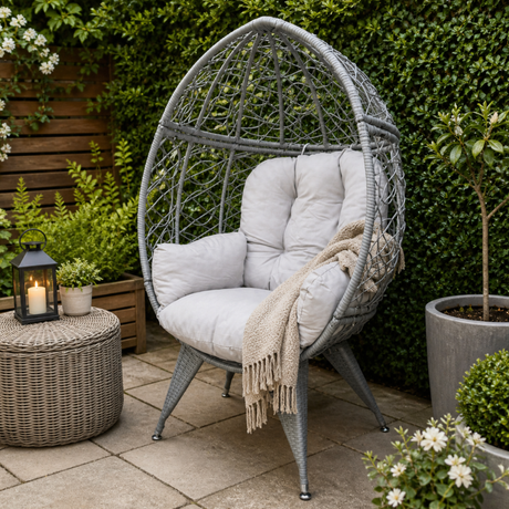 Gray wicker egg-shaped chair with white cushions on a patio, surrounded by plants and a lantern.