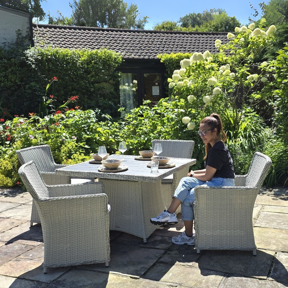 Woman sitting at a patio table in a garden setting with greenery and flowers.
