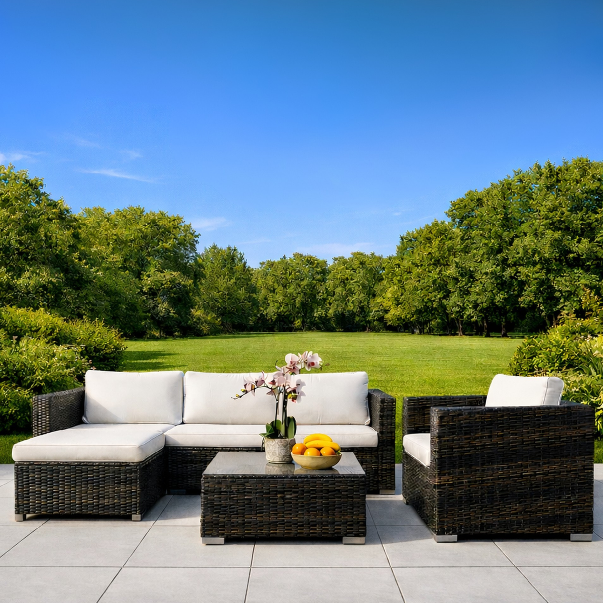 Outdoor patio set with white cushions and a small table with flowers and fruit on a patio with a green lawn and trees in the background.