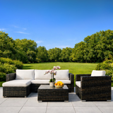 Outdoor patio set with white cushions and a small table with flowers and fruit on a patio with a green lawn and trees in the background.