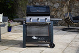 A black Sahara S375 3 Burner Gas BBQ with side burner, placed on a tiled outdoor area with a rock wall and a chair in the background.