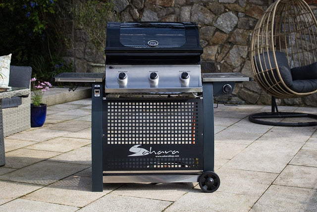 A black Sahara S375 3 Burner Gas BBQ with side burner, placed on a tiled outdoor area with a rock wall and a chair in the background.
